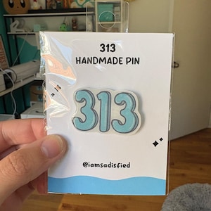 May include: A handmade pin featuring the number "313" in a light blue color with a white outline. The pin is on a white card with the text "313 HANDMADE PIN" and the Instagram handle "@iamsatisfied".
