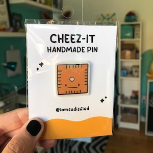 May include: A white card with the text "CHEEZ-IT HANDMADE PIN" and a cartoon image of a Cheez-It cracker with a face. The card also includes the text "@iamsatisfied".