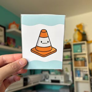 May include: A small, rectangular card featuring a cartoon traffic cone with a minimalist face. The cone is orange with a white band and sits on a square base. The background has a light blue wavy design. The card is held in a hand.