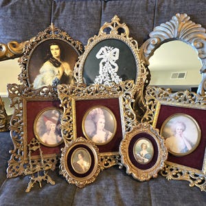 May include: A collection of ornate gold-toned frames and mirrors, some featuring oval portraits of individuals in period attire, others containing a white lace bow or reflective glass, all arranged together with intricate scrollwork and velvet accents.