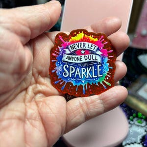 May include: A colorful sticker with the text "NEVER LET ANYONE DULL YOUR SPARKLE." The sticker has a red glittery border and features a vibrant design with blue, pink, yellow, and green accents. The text is in a stylized font.