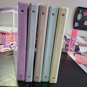 May include: A row of five pastel-colored binders. The binders are in shades of lavender, light gray, beige, seafoam green, and pale yellow. Each binder has a metal ring mechanism and a button closure. The binders are standing upright on a dark surface.