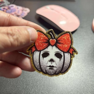 May include: A Halloween-themed pin featuring a pumpkin design with a bow. The pumpkin has a face resembling a horror movie character. The bow is red with glitter, and the pin has a gold glitter border.