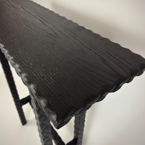 May include: A black wooden console table with a textured, wavy edge. The tabletop has a wood grain pattern. The legs are made of a dark, textured material. The table is approximately 48 inches long.