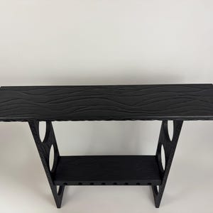 May include: Black console table with a textured, wave-like pattern on the tabletop. The table has a lower shelf and angled legs with circular cutouts. The table is approximately 48 inches long.
