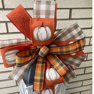 May include: A decorative bow made of multiple ribbons featuring orange fabric with white pumpkin illustrations, brown and white buffalo check patterns, and a multi-colored plaid design with orange, yellow, and dark blue stripes throughout the loops.