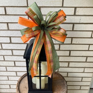 May include: A decorative bow featuring layers of orange, olive green, and plaid ribbon with white polka dots, attached to the top of a black metal lantern containing three white pillar candles. The bow has multiple loops and long, flowing tails.