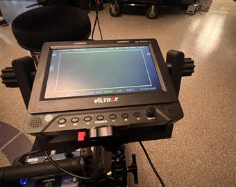 Puede incluir: Un monitor Viltrox negro con una pantalla que muestra una imagen verde. El monitor tiene varios botones y una perilla en la parte delantera. El monitor está montado en un equipo de cámara.