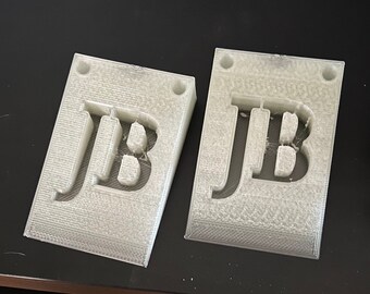 Puede incluir: Dos topes de puerta de plástico blanco con las iniciales "JB" recortadas en el centro. Los topes de puerta son rectangulares y tienen una superficie texturizada.