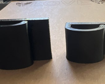 Puede incluir: Dos soportes de pared impresos en 3D de color negro con un diseño curvo. Los soportes están diseñados para sujetar objetos como un teléfono o una tableta.