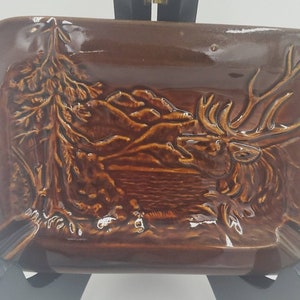 May include: Brown ceramic ashtray with a raised relief design. The design features a majestic elk, mountains, a lake, and a tree. The ashtray has a rectangular shape with rounded corners and a glossy finish.