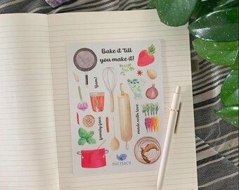 May include: An open notebook with lined pages, featuring a sticker sheet with watercolor illustrations of cooking tools and ingredients. The sticker sheet includes text that says "Bake it 'till you make it!" and "family fave". A white pen rests on the notebook.