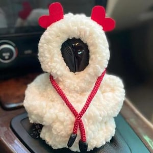 May include: A white, fluffy car accessory shaped like a sheep, with red heart-shaped accents and a red cord. The sheep-shaped item is placed on a car's gear shift.