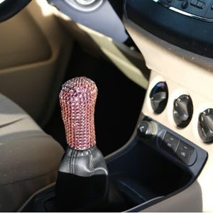 Op de afbeelding: Een roze strass versnellingspookknop voor een auto. De knop is gemaakt van een zwarte lederen basis met een roze strass top.
