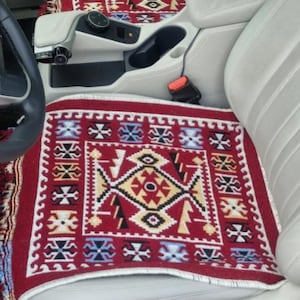 Może przedstawiać: Czerwone i kremowe pokrowce na siedzenia samochodowe z geometrycznym wzorem. Pokrowce są umieszczone na przednich siedzeniach samochodu. Wzór zawiera czarne, żółte i niebieskie akcenty z frędzlami.