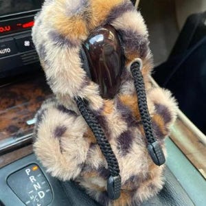 Pode incluir: Uma capa de alavanca de câmbio de carro com estampa de leopardo. A capa é marrom, bege e preta, com cordões e batentes pretos. É projetada para caber na alavanca de câmbio de um veículo.