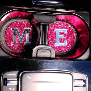 Könnte beinhalten: Einsätze für Getränkehalter im Auto mit rosa Glitzerdesign. Die Einsätze zeigen die Buchstaben "M" und "E" in Silber und Hellblau. Die Konsole des Autos ist schwarz und silber.