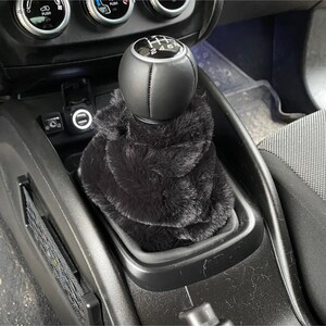 May include: Black faux fur gear shift boot cover for a car. The cover is made of soft, plush faux fur and fits over the gear shift knob. The gear shift knob is black leather with a silver gear shift pattern.
