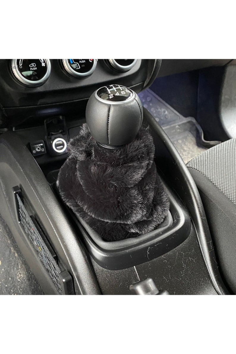 May include: Black faux fur gear shift boot cover for a car. The cover is made of soft, plush faux fur and fits over the gear shift knob. The gear shift knob is black leather with a silver gear shift pattern.