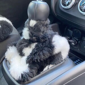 May include: Black and white faux fur gear shift cover for a car. The cover is made of soft, fluffy material and has a black leather gear knob.
