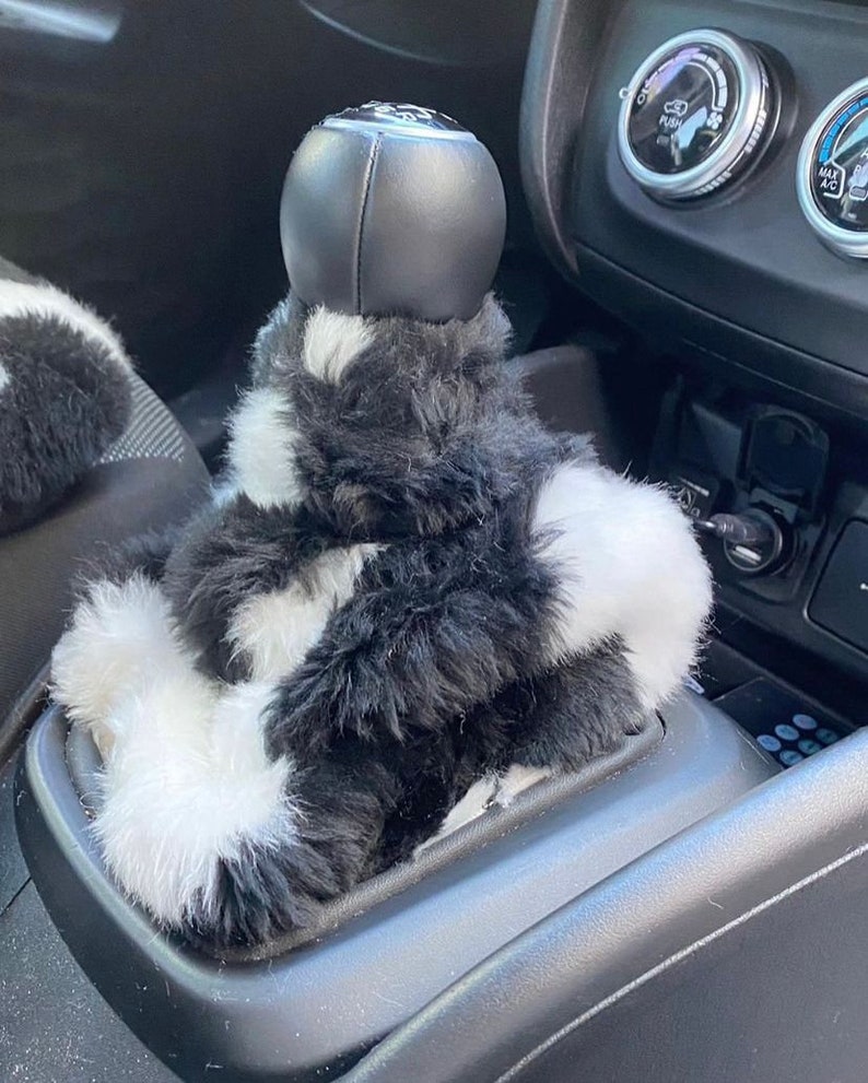 May include: Black and white faux fur gear shift cover for a car. The cover is made of soft, fluffy material and has a black leather gear knob.