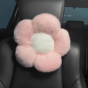 May include: A plush, flower-shaped pillow in soft pink and white. The pillow is designed with a central white circle surrounded by pink petals. It is placed on a black car seat, suggesting its use as a decorative or supportive accessory.