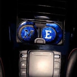 May include: Two blue glitter car cup holders with the letters "M" and "E" in silver on top. The cup holders are in a car's centre console next to a black control panel with buttons for dynamic, volume, navi map, radio media, and tel.