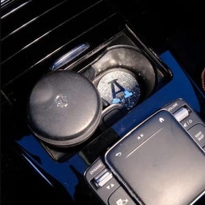 Pode incluir: Um porta-copos de carro preto com um porta-copos de glitter prata e azul com a letra "A". O porta-copos está no console central do carro. O console tem botões para "Dynamic", "Navi Map" e "Volume".