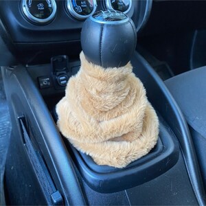 May include: A tan faux fur gear shift cover for a car. The cover is made of soft, plush fabric and is designed to fit over a standard gear shift knob.