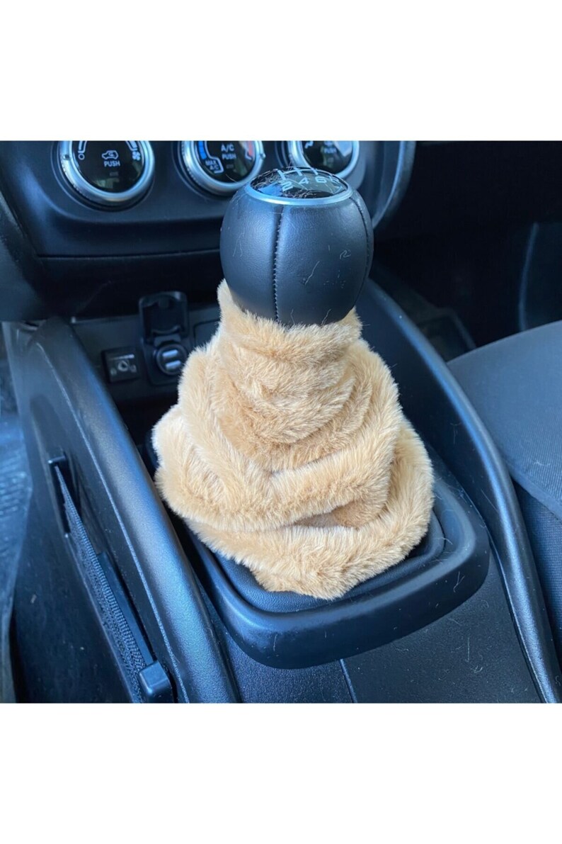 May include: A tan faux fur gear shift cover for a car. The cover is made of soft, plush fabric and is designed to fit over a standard gear shift knob.