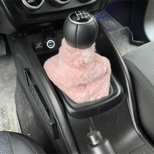 May include: Pink faux fur gear shift cover for a car. The cover is over a black leather gear shift knob with a silver gear shift pattern that says "1 3 5 R 2 4 6".