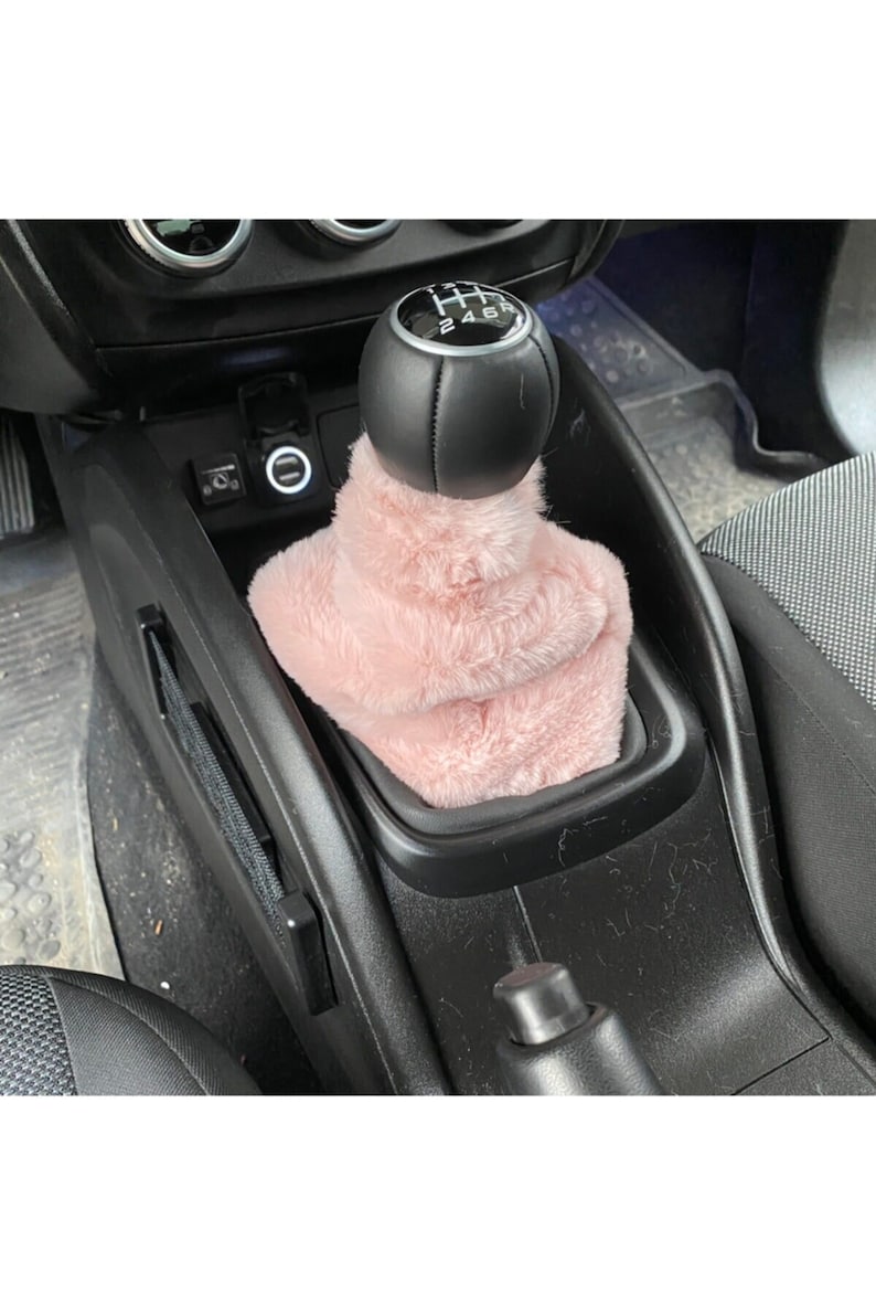 May include: Pink faux fur gear shift cover for a car. The cover is over a black leather gear shift knob with a silver gear shift pattern that says "1 3 5 R 2 4 6".