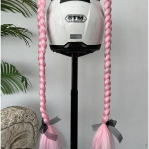Könnte beinhalten: Ein weißer Motorradhelm mit schwarzen Akzenten und rosa geflochtenen Haarverlängerungen. Die Verlängerungen sind mit schwarzen und grauen Bändern verziert. Der Helm ist auf einem schwarzen Ständer montiert.