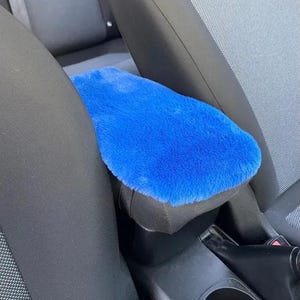 Può includere: Una copertura per bracciolo per auto blu brillante e soffice. La copertura è rettangolare e si adatta al bracciolo della console centrale. L'interno dell'auto circostante è nero e grigio.