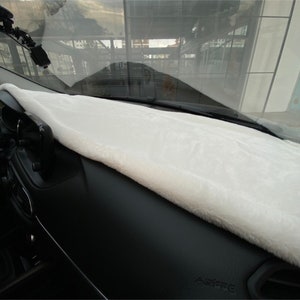 Könnte beinhalten: Weiße Kunstfell-Armaturenbrett-Abdeckung für ein Auto. Die Abdeckung wird auf dem Armaturenbrett platziert und erstreckt sich bis zur Windschutzscheibe. Die Abdeckung besteht aus weichem, plüschigem Material und ist so konzipiert, dass sie das Armaturenbrett vor Sonnenschäden und Kratzern schützt.