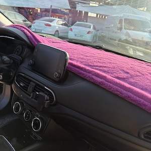 Könnte beinhalten: Auto-Armaturenbrett mit einer leuchtend rosa, flauschigen Abdeckung. Das schwarze Armaturenbrett hat einen zentralen Bildschirm und Lüftungsschlitze. Die Windschutzscheibe zeigt geparkte Autos und ein Gebäude.