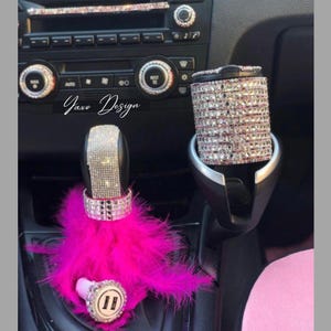 Peut inclure: Un pommeau de levier de vitesse de voiture en plumes roses avec un dessus recouvert de cristaux et un porte-gobelet assorti recouvert de cristaux. Le texte "Yaxo Design" est visible sur l'image.