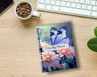 May include: A spiral-bound monthly planner featuring a large butterfly with blue and purple wings, and pink flowers. The words "Monthly Planner" are printed on the cover. A cup of coffee beans and a keyboard are also visible.