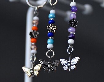 May include: Three keychains with butterfly charms and colorful beads. The keychains feature a silver butterfly charm, a gold butterfly charm, and a silver butterfly charm with purple, white, and blue beads.
