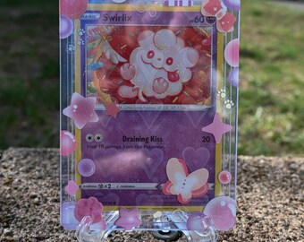 May include: A Swirlix Pokémon card in a clear protective case, displayed on a clear stand. The card features a pink and white cartoon character with the name "Swirlix" at the top. The case is decorated with pink stars and bubbles.