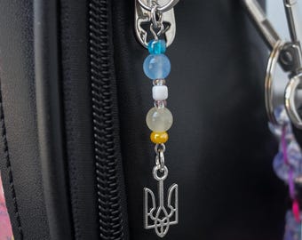 May include: A silver-toned metal charm featuring the Ukrainian Tryzub symbol, attached to a black bag zipper pull. The charm is suspended from a vertical strand of small, multi-colored beads in shades of blue, white, yellow, and translucent clear.