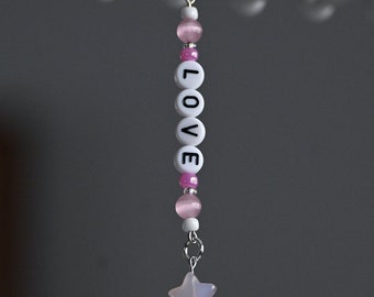 May include: A white and pink beaded keychain with the word "LOVE" spelled out in black letters. The keychain has a small, light pink star charm at the bottom.