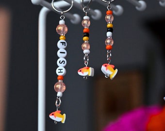 May include: Three beaded keychains with a small fish charm. The keychain on the left has white beads with the word "FISH" spelled out in black letters. The other two keychains have orange, white, and black beads.
