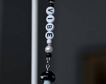 May include: A black and white beaded keychain with the word "VIBE" spelled out on white beads. The keychain has a black and white mushroom charm at the end.