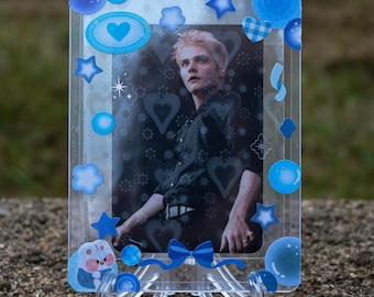 May include: A clear plastic card holder with a photo of a person with blonde hair. The holder is decorated with blue stickers, including stars, hearts, and a bow. The card holder is displayed on a clear plastic stand.