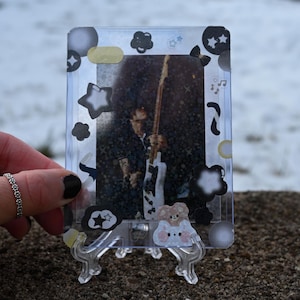 Puede incluir: Un tarjetero de plástico transparente con pegatinas decorativas con estrellas, nubes y un oso. El soporte muestra una foto de una persona tocando la guitarra. El soporte está sostenido por un pequeño soporte de plástico transparente.
