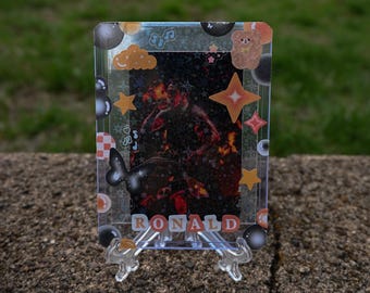 May include: A clear, rectangular card holder with decorative stickers and the name "RONALD" in red letters. The card inside features a dark image with red and orange hues, and is decorated with stars, clouds, and a butterfly.