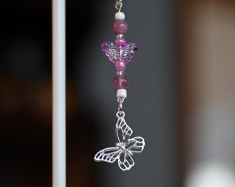 May include: A silver butterfly charm with pink and white beads hangs from a black phone strap.