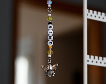 May include: A white and yellow beaded keychain with the words "BEE KIND" spelled out in black letters. A silver bee charm hangs from the bottom of the keychain.