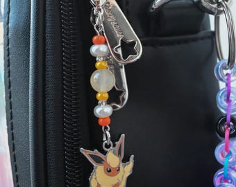 May include: A bag charm attached to a black zipper pull, featuring a metal Flareon character pendant. The charm includes a vertical arrangement of small orange, white, and yellow beads, along with two white pearls, connected by a silver-toned metal chain.